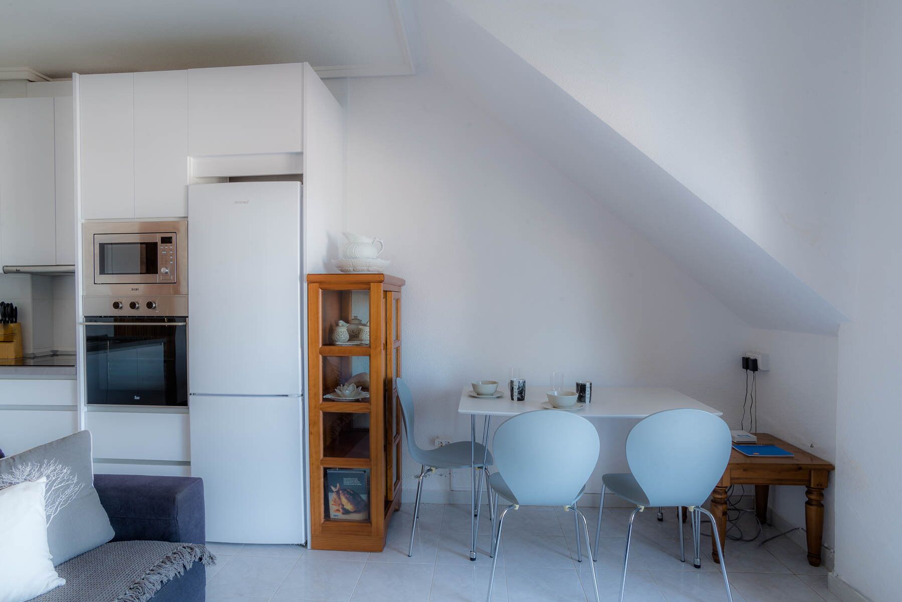 Une cuisine compacte équipée d’un réfrigérateur, d’un four et d’un four à micro-ondes. On y trouve une table à manger avec deux chaises et un petit meuble contenant des objets décoratifs.
