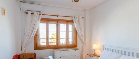 Une chambre à coucher comprenant un lit, une table de chevet, une commode, une fenêtre avec des rideaux, un ventilateur de plafond et un petit tabouret en osier.