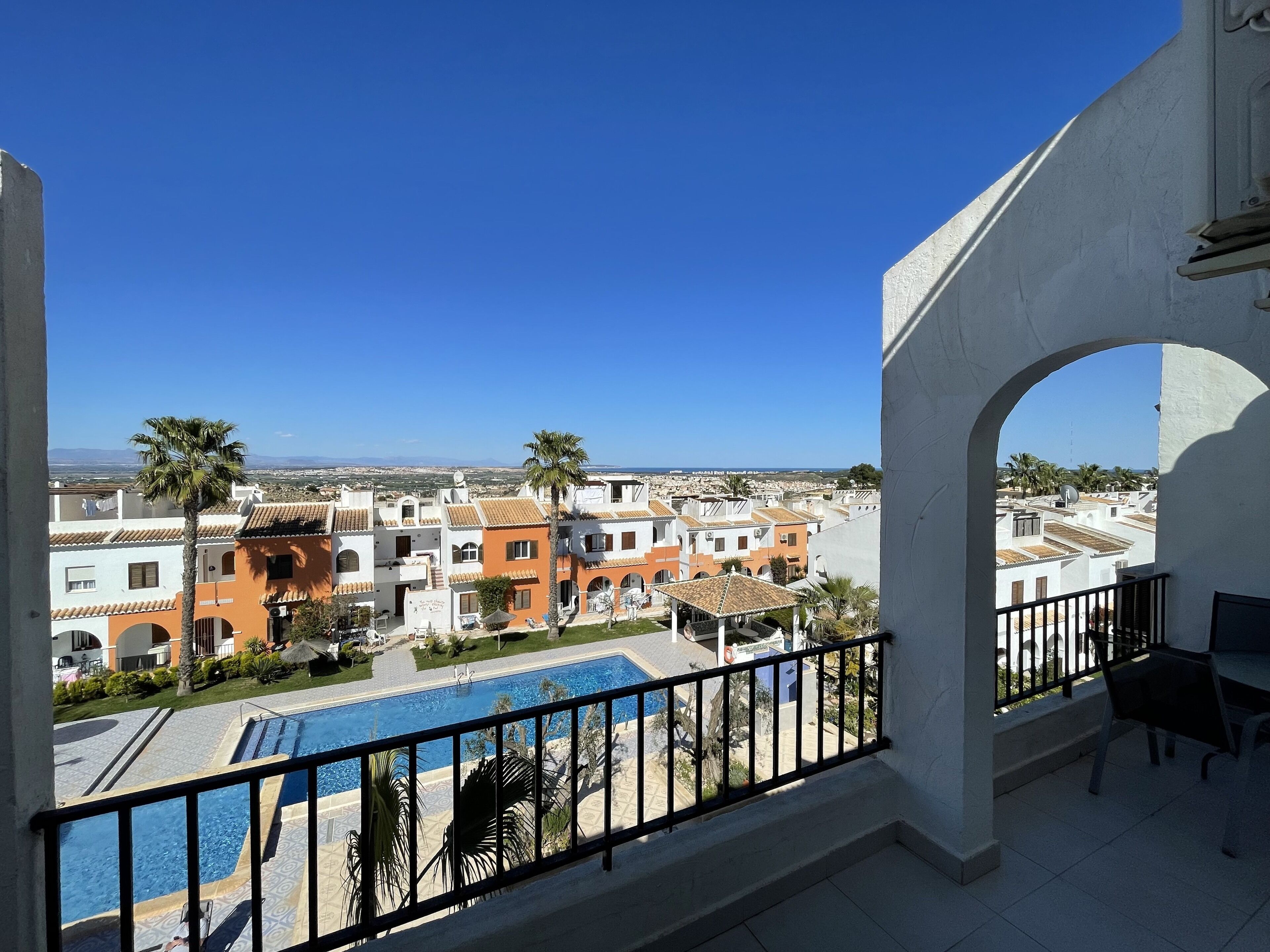 A balcony with a view of a swimming pool, a palm tree, and buildings in the background.