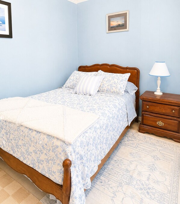 A bedroom with a wooden bed, a nightstand with a lamp, a framed picture on the wall, and a patterned rug. - Castaway Cottage in Sylvan Beach (Sylvan Beach)