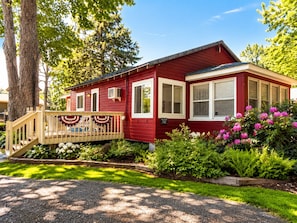 A red house with a porch, a deck, and a garden with pink flowers.