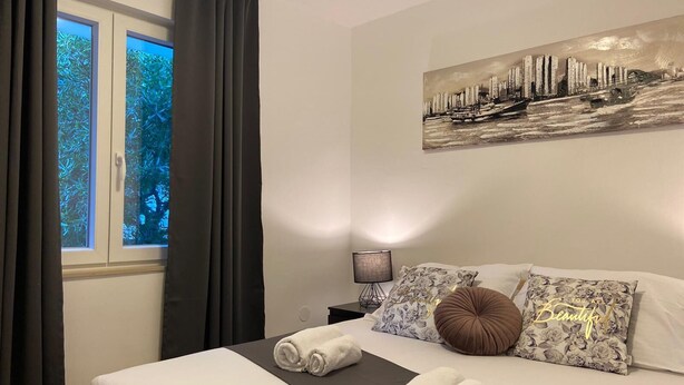 Un lit bien fait, avec des serviettes pliées, un tableau représentant un paysage urbain accroché au mur et une fenêtre avec des rideaux.