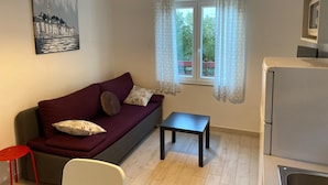 Un espace de vie compact comprenant un canapé, une petite table, un tabouret rouge, un coin cuisine avec un réfrigérateur et une fenêtre avec des rideaux.