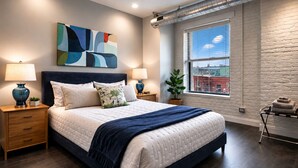 Un dormitorio moderno con una cama grande, dos mesitas de noche con lámparas, un cuadro abstracto en la pared, una ventana con vistas a la ciudad y una pared de ladrillo.