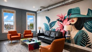 Un salón moderno con dos sillones naranjas, un sofá de cuero negro, una mesa de centro colorida y un gran mural tropical.