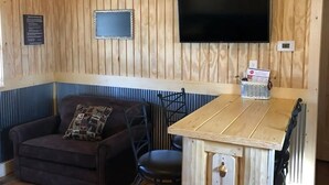 Una habitación con paneles de madera, un televisor de pantalla plana en la pared, una mesita con una bebida y un sofá con un cojín estampado.