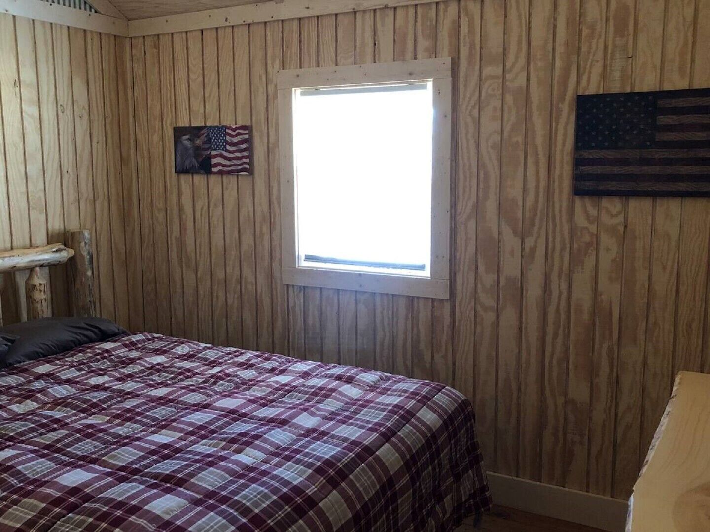 Quarto com paredes de madeira, uma janela, uma cama com colcha xadrez e uma bandeira dos Estados Unidos na parede.