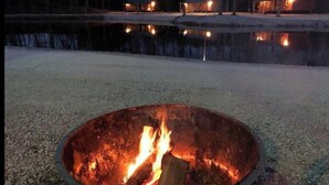En eldstad med flammor, omgiven av ett område med grus och en vattenyta i bakgrunden.