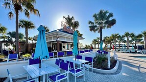 An outdoor seating area with white tables and blue chairs, surrounded by palm trees and a pool.