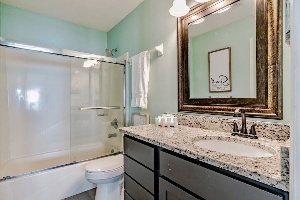 A bathroom with a glass shower enclosure, a white toilet, a granite countertop sink, and a large mirror with a decorative frame.