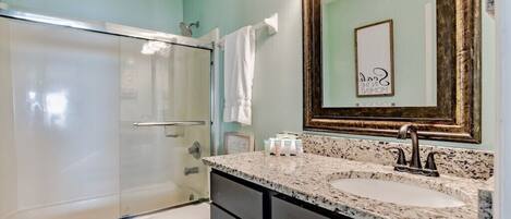 A bathroom with a glass shower enclosure, a white toilet, a granite countertop sink, and a large mirror with a decorative frame.