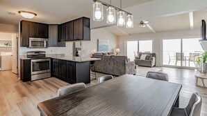 Una cucina moderna con mobili scuri, elettrodomestici in acciaio inossidabile e un'isola centrale. C'è una zona pranzo con un tavolo grande e sedie. La stanza ha un soffitto alto con ventilatore a soffitto e luci a sospensione.