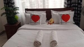 Una cama bien tendida con ropa de cama blanca, almohadones con forma de corazón, un pequeño peluche y un ventilador de pie.