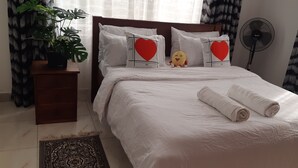 Una cama bien hecha con ropa de cama blanca, una planta en maceta y almohadones decorativos con diseños de corazones.