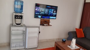 Sala de estar con televisor de pantalla plana montado en la pared, dispensador de agua, minibar, sofá con cojines anaranjados y una mesa de centro con un control remoto y un objeto decorativo.