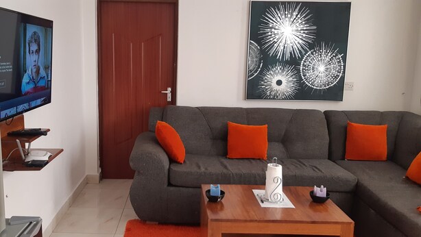 Una sala de estar con un sofá gris, cojines anaranjados, una mesa de centro, un dispensador de agua y un televisor mostrando una serie de Netflix.