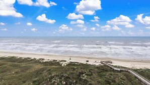 Ein Strand mit einer hölzernen Promenade, die zu einem Sandbereich führt, einem grasbewachsen Vordergrund und einem strahlend blauen Himmel mit Wolken.