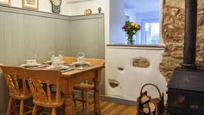 A dining area with a wooden table and chairs, a stone wall, a window with a view, and a black wood stove. - 2 bedroom accommodation in Clapham, near Ingleton (Clapham, near Ingleton)