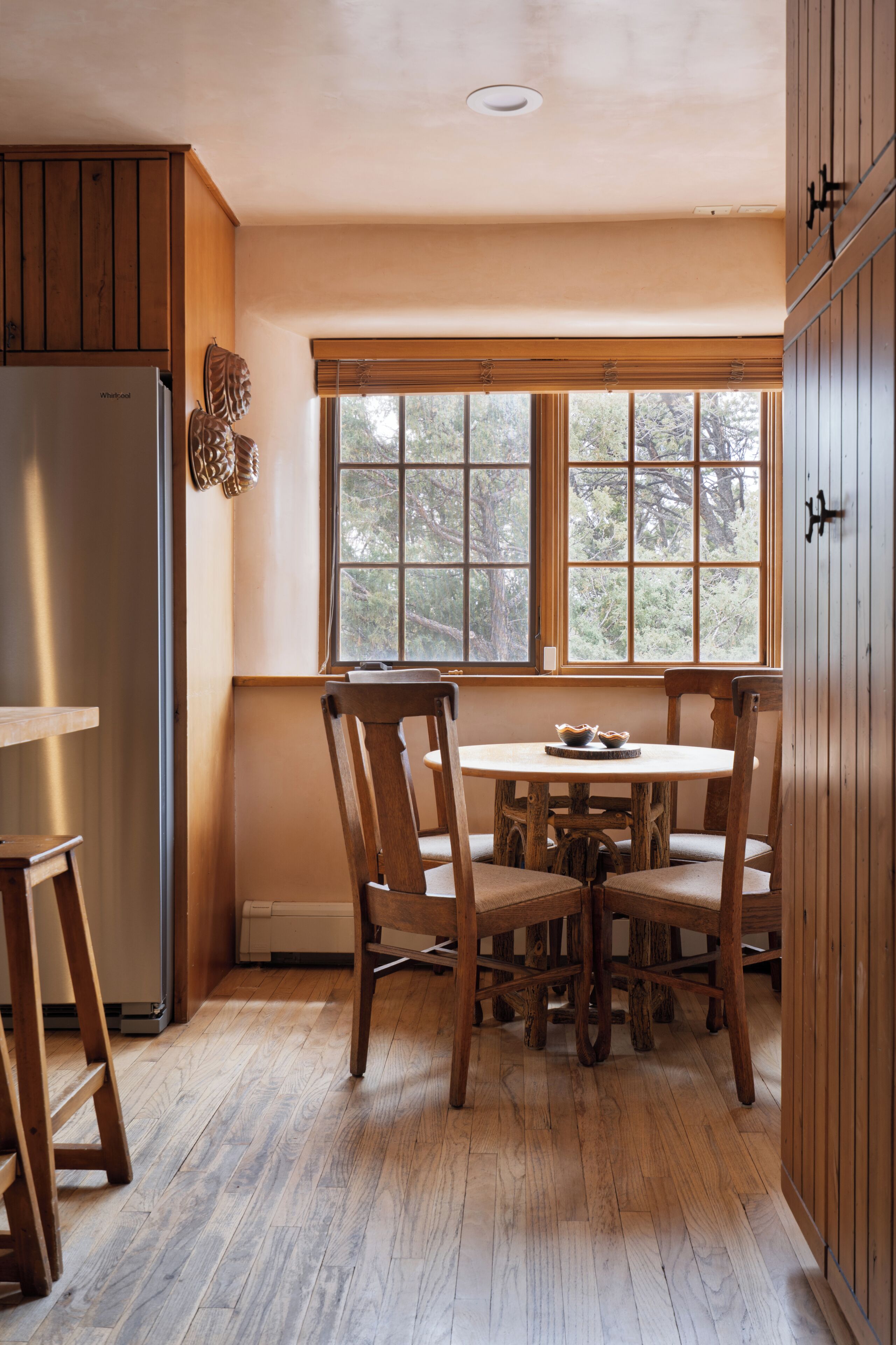 A kitchen with wooden cabinets, a refrigerator, a dining table with chairs, and a window with a view of trees.