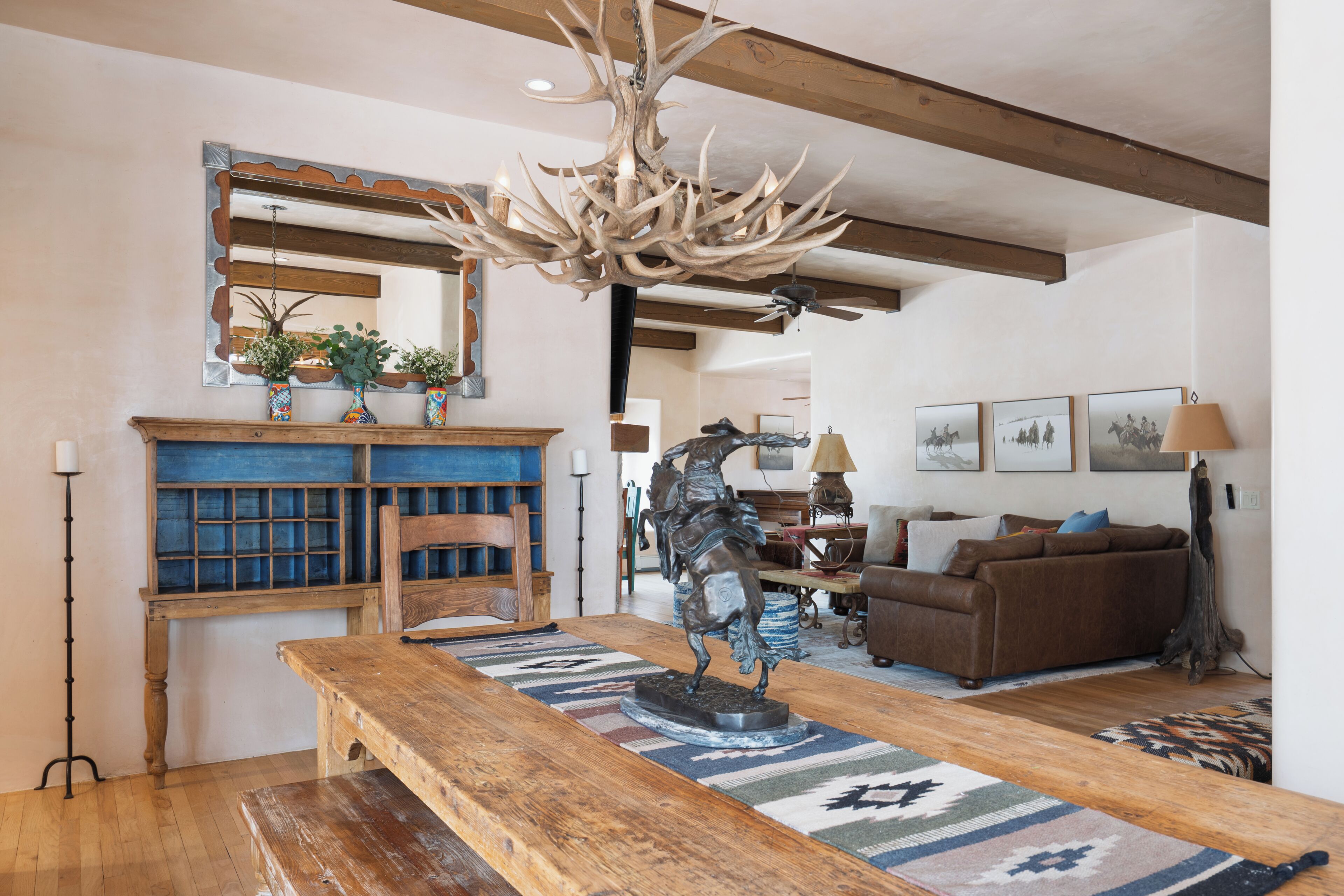 A rustic interior with a wooden dining table, a chandelier with antlers, a fireplace, and a sofa.