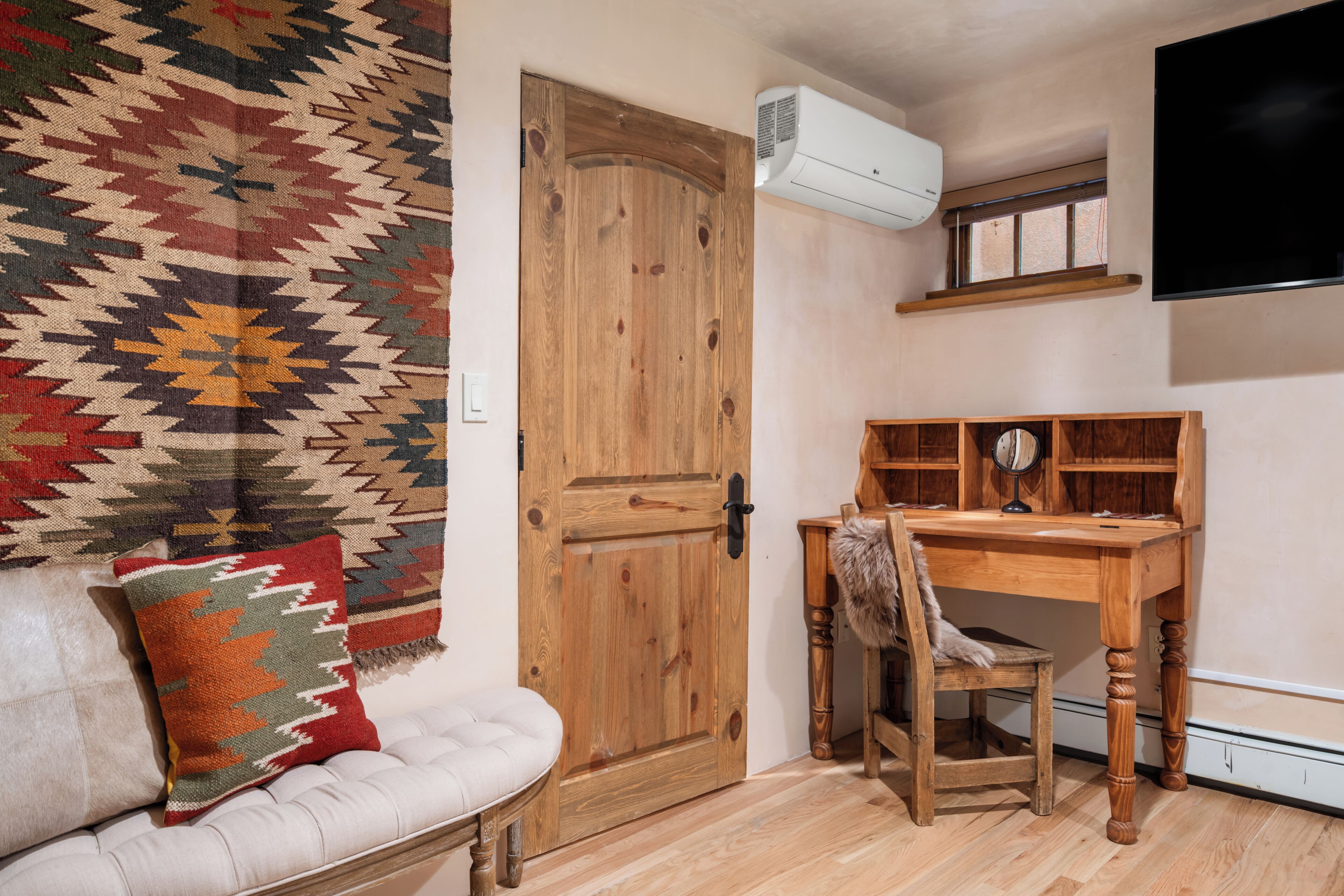 A room with a wooden door, a white cushioned bench, a wooden desk with a chair, and a mounted television.
