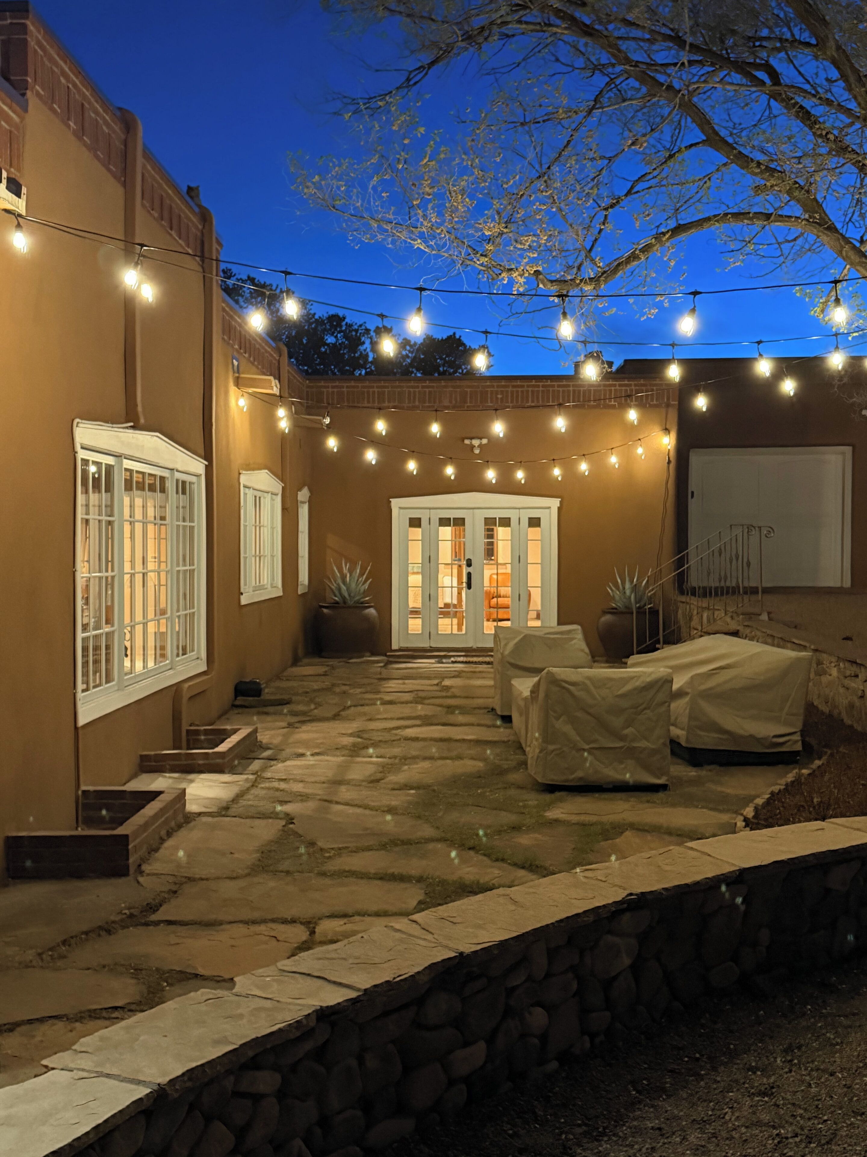 Un patio con suelo de piedra, mobiliario cubierto y luces colgantes.