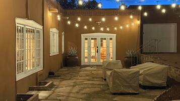 Un patio con suelo de piedra, mobiliario cubierto y luces colgantes.