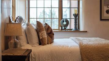 Una cama bien hecha con colcha blanca, una lámpara en una mesita de noche y una ventana con persianas.