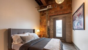 A bedroom with a bed, a nightstand, a lamp, a door, and a brick wall. - Luxury Downtown Executive Loft @ the ABODES ApartHotel (Rochester)