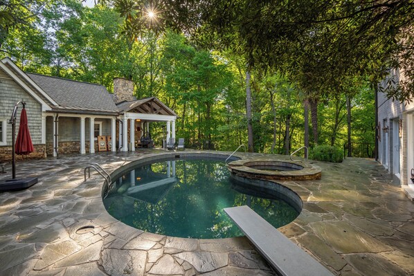 A stone pool with a hot tub, surrounded by a covered patio and a stone house.