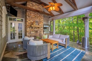 A covered outdoor seating area with stone walls, a grill, and a view of a forest.