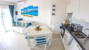Een lichte, moderne keuken met witte kastjes, een roestvrijstalen spoelbak en een magnetron. Er is een eethoek met een witte tafel en blauwe stoelen, en een bank met kleurrijke kussens.