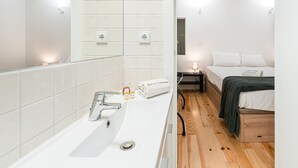 Un bany modern amb un rentapans blanc, mirall i parets revestides de rajola. Hi ha un paviment de fusta, un llit amb roba de llit blanca i una taula de nit amb una làmpada.