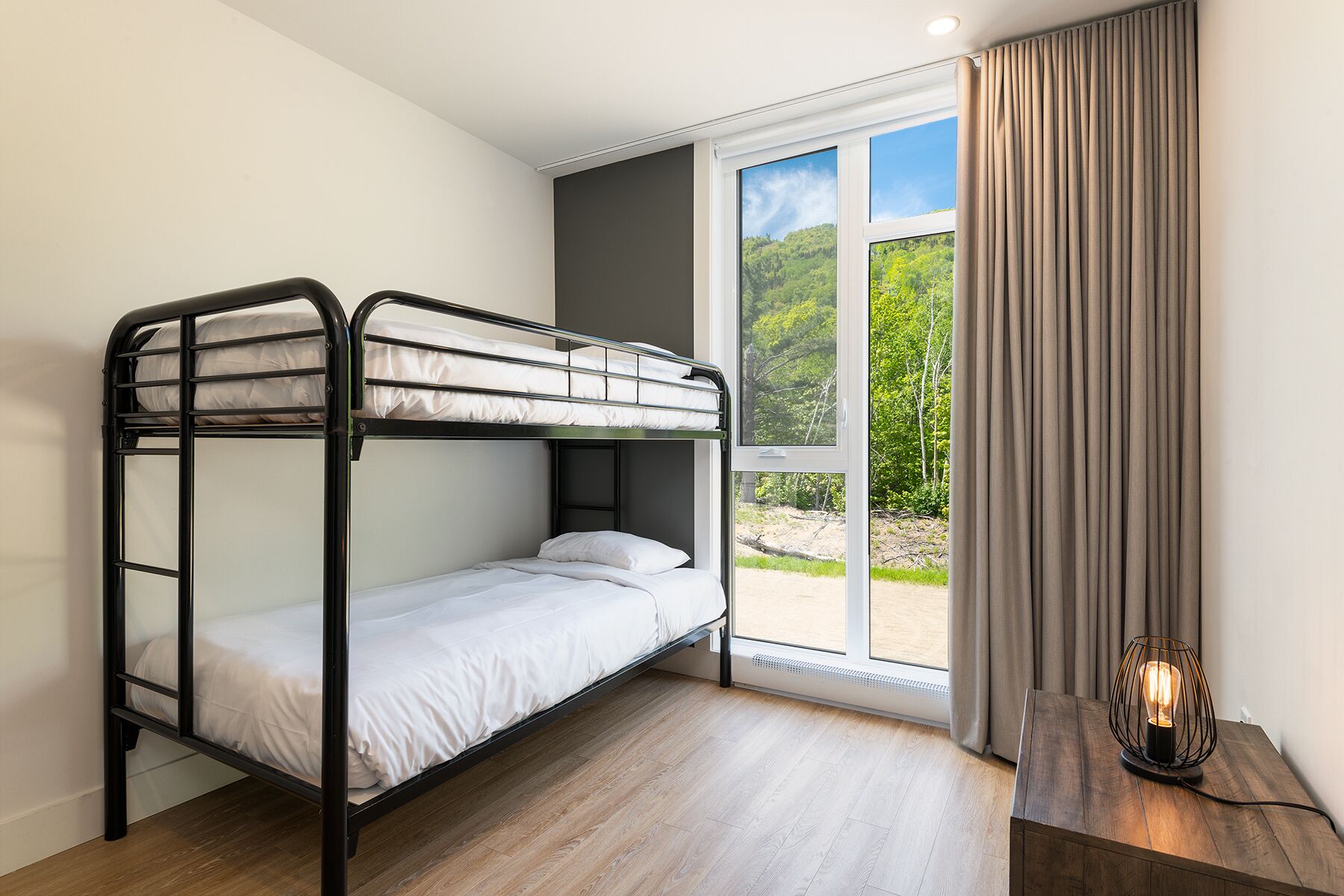 A bunk bed room with a window and curtains, a wooden floor, and a table with a lamp.