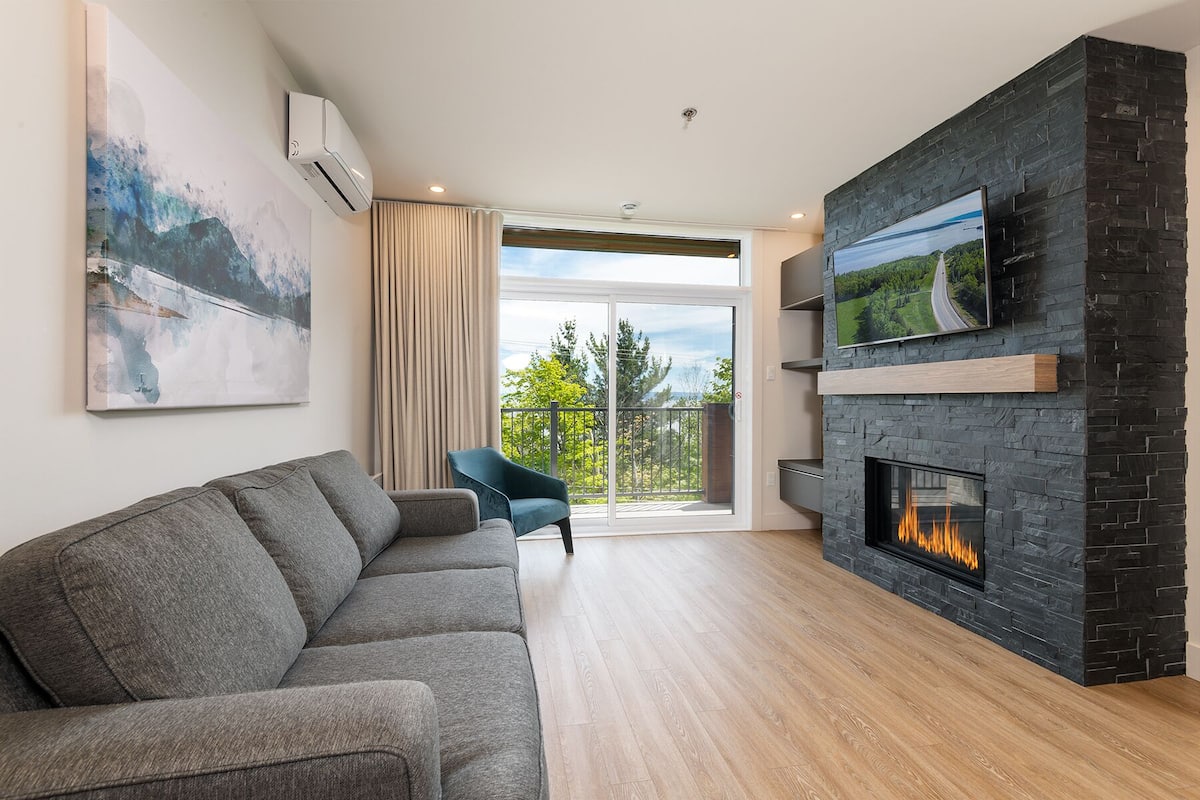 A modern living room with a grey sofa, a fireplace, a TV mounted on a stone wall, and a large window with a view of trees and a road.