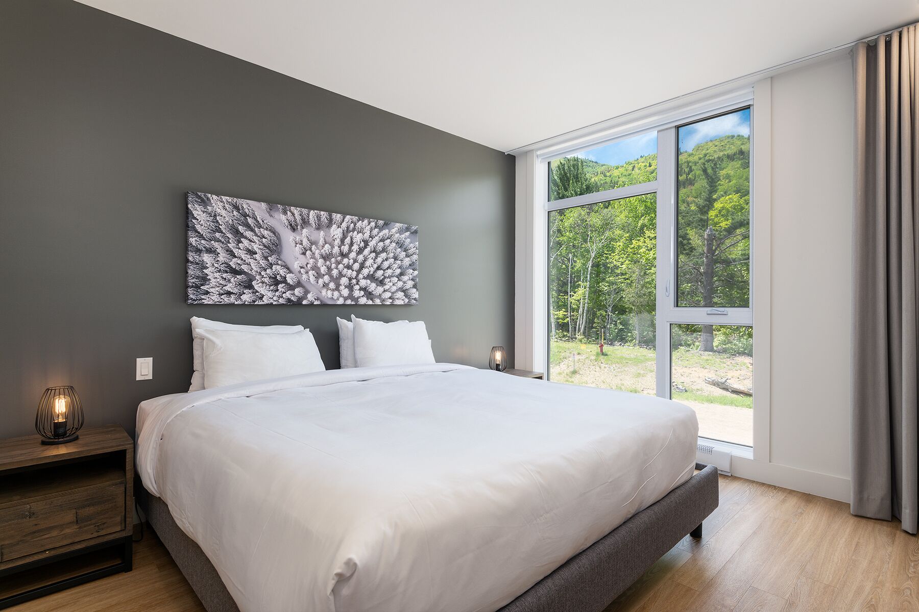 Une chambre moderne avec un grand lit, une table de chevet, une lampe fixée au mur et une fenêtre donnant sur des arbres.
