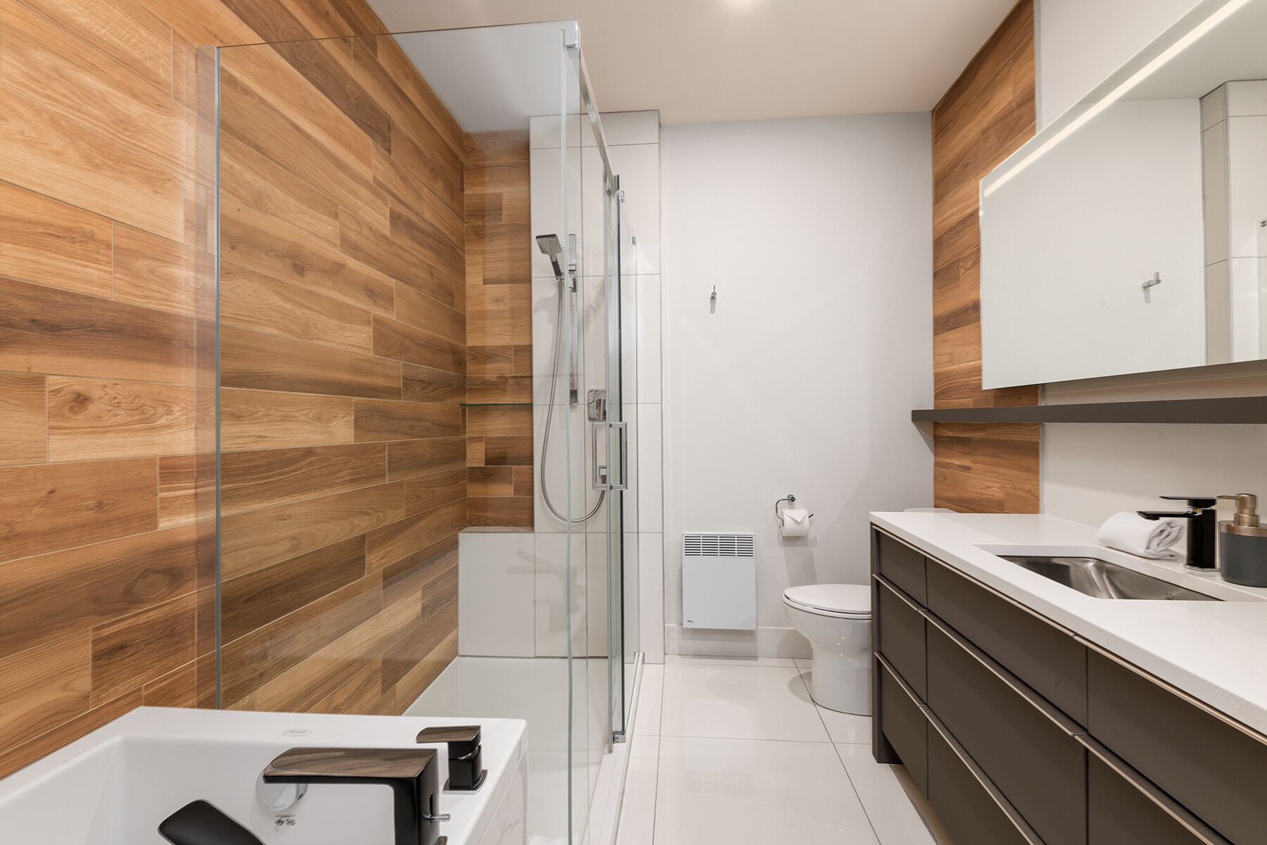 A modern bathroom with wooden walls, a glass shower enclosure, a white toilet, and a dark-toned vanity with a mirror.