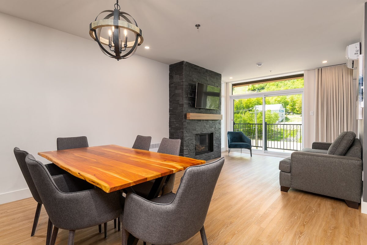 A modern living room with a wooden dining table, grey upholstered chairs, a stone fireplace, and a large window offering a view of greenery.