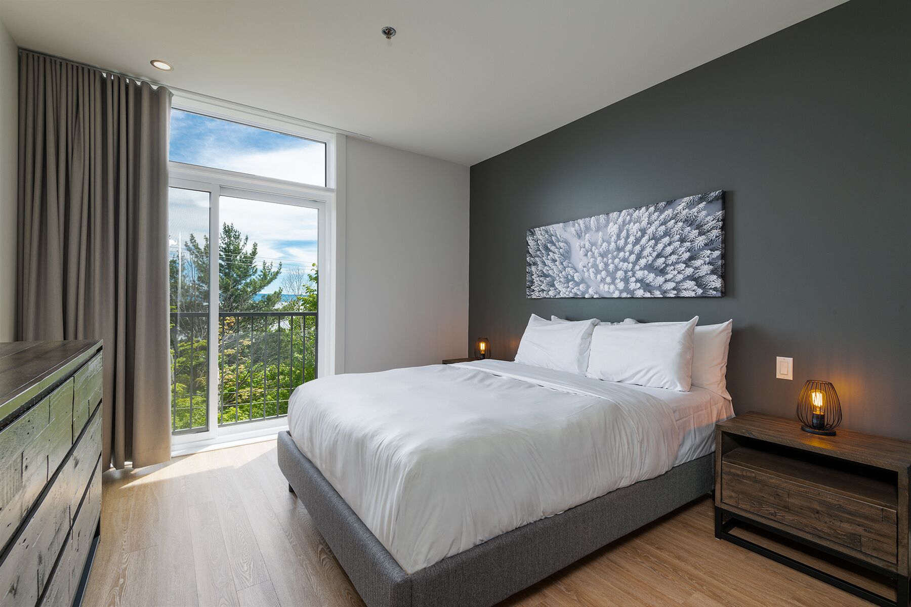 Une chambre moderne avec un grand lit, une vue sur la verdure et un mur foncé orné d’une œuvre d’art à motifs de feuilles.