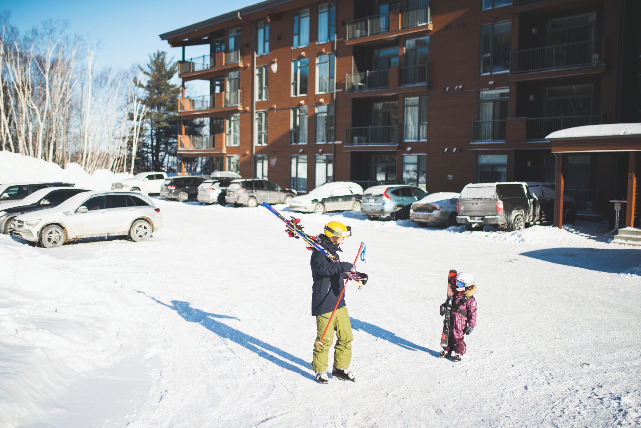 A building with multiple floors and balconies, surrounded by parked cars and snow on the ground.