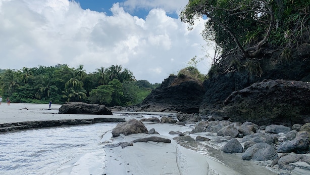 Una spiaggia con grandi rocce, acqua calma e palme.
