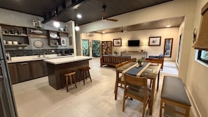 Una cucina moderna con isola centrale, sgabelli da bar, zona pranzo con tavolo in vetro e soggiorno con divano e televisore.