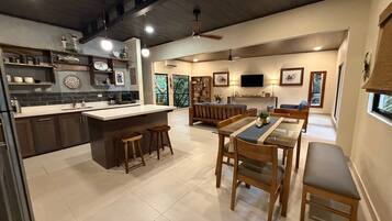 Une cuisine moderne dotée d’un îlot central, de tabourets de bar, d’un coin repas avec une table en verre et d’un salon avec un canapé et une télévision.
