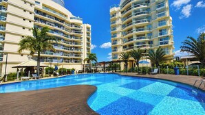 Una piscina con muelle de madera, rodeada por dos edificios altos con balcones, palmeras y sillas de descanso.