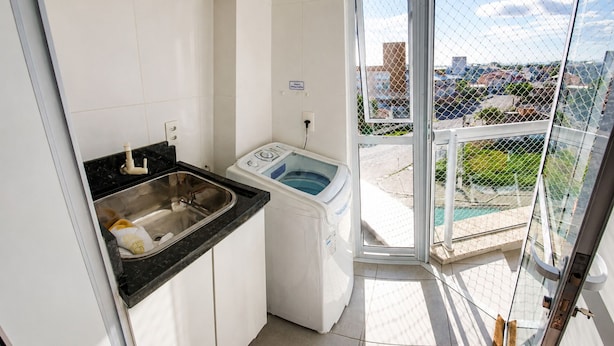 Une petite cuisine équipée d’un évier et d’un lave-linge, un balcon avec vue sur la ville et une porte vitrée donnant sur l’extérieur.
