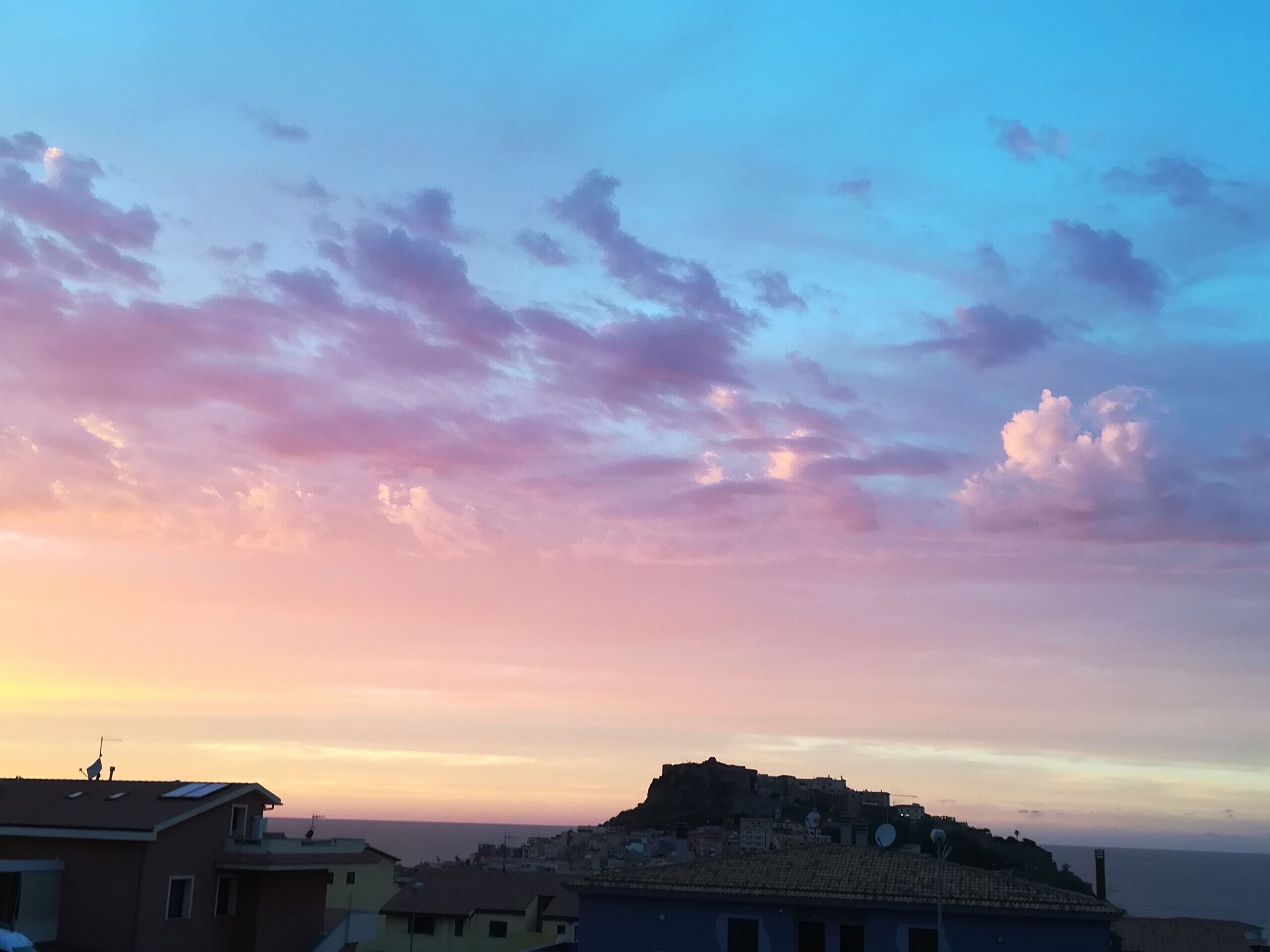 A coastal cityscape with a prominent hilltop fortress, buildings with flat roofs, and a clear sky with scattered clouds.