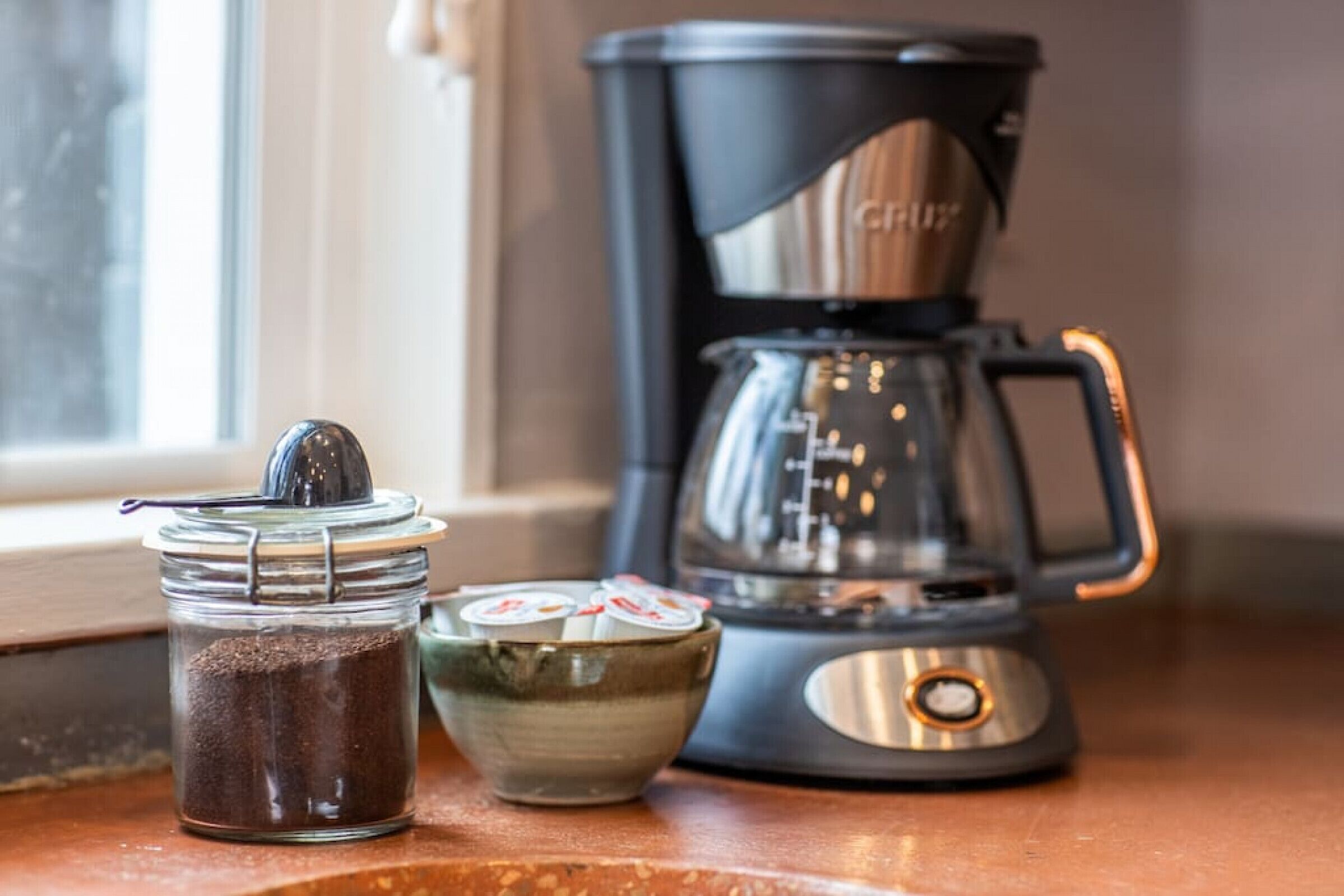 A coffee maker, a jar of coffee grounds, and a small bowl with coffee pods on a kitchen counter.