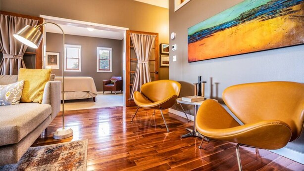 Una habitación de hotel moderna con suelo de madera, un sofá, dos sillones amarillos, un cuadro en la pared y una cama al fondo.