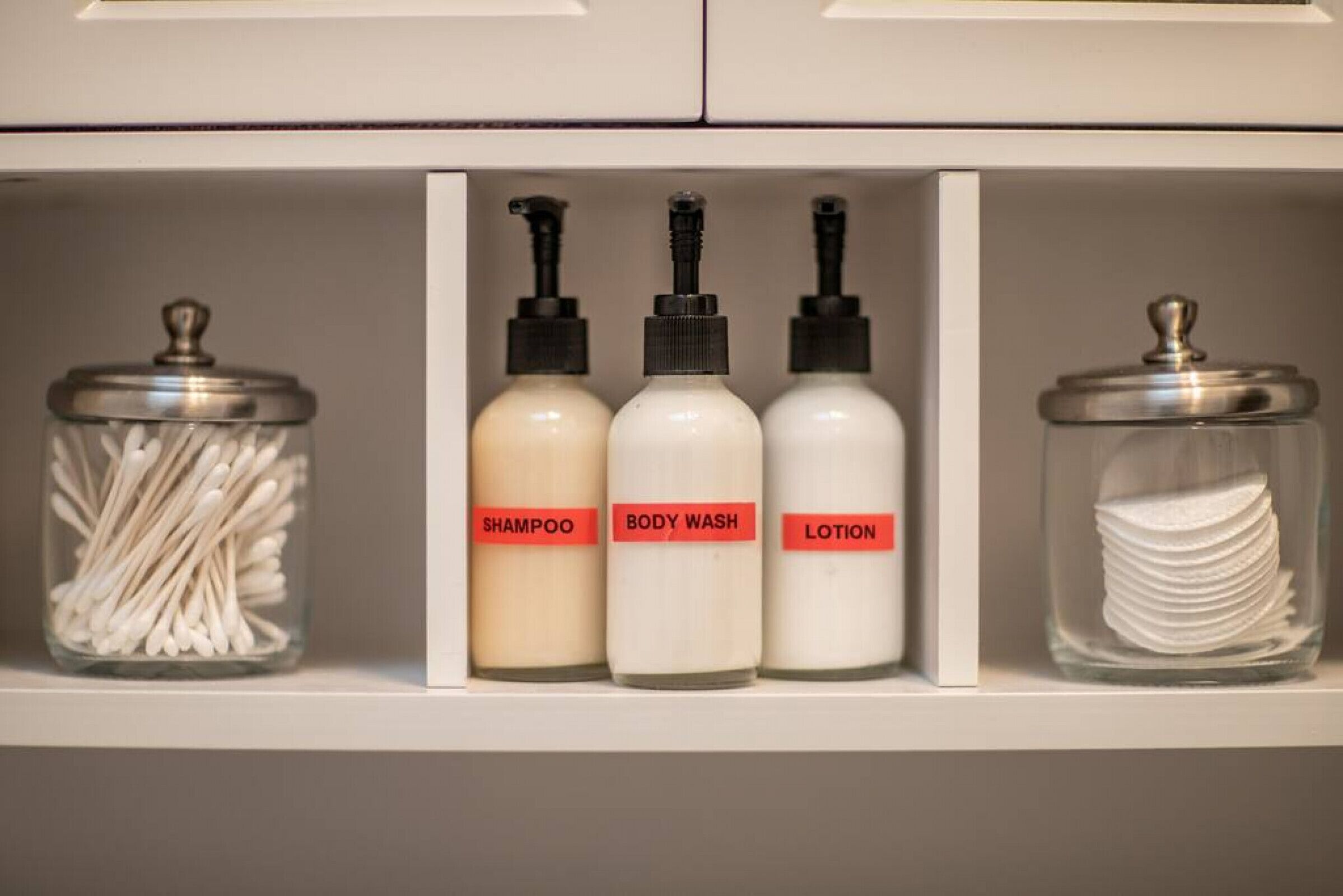 A bathroom shelf with three bottles labeled shampoo, body wash, and lotion, and a jar of cotton swabs.