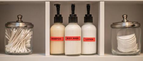 A bathroom shelf with three bottles labeled shampoo, body wash, and lotion, and a jar of cotton swabs.